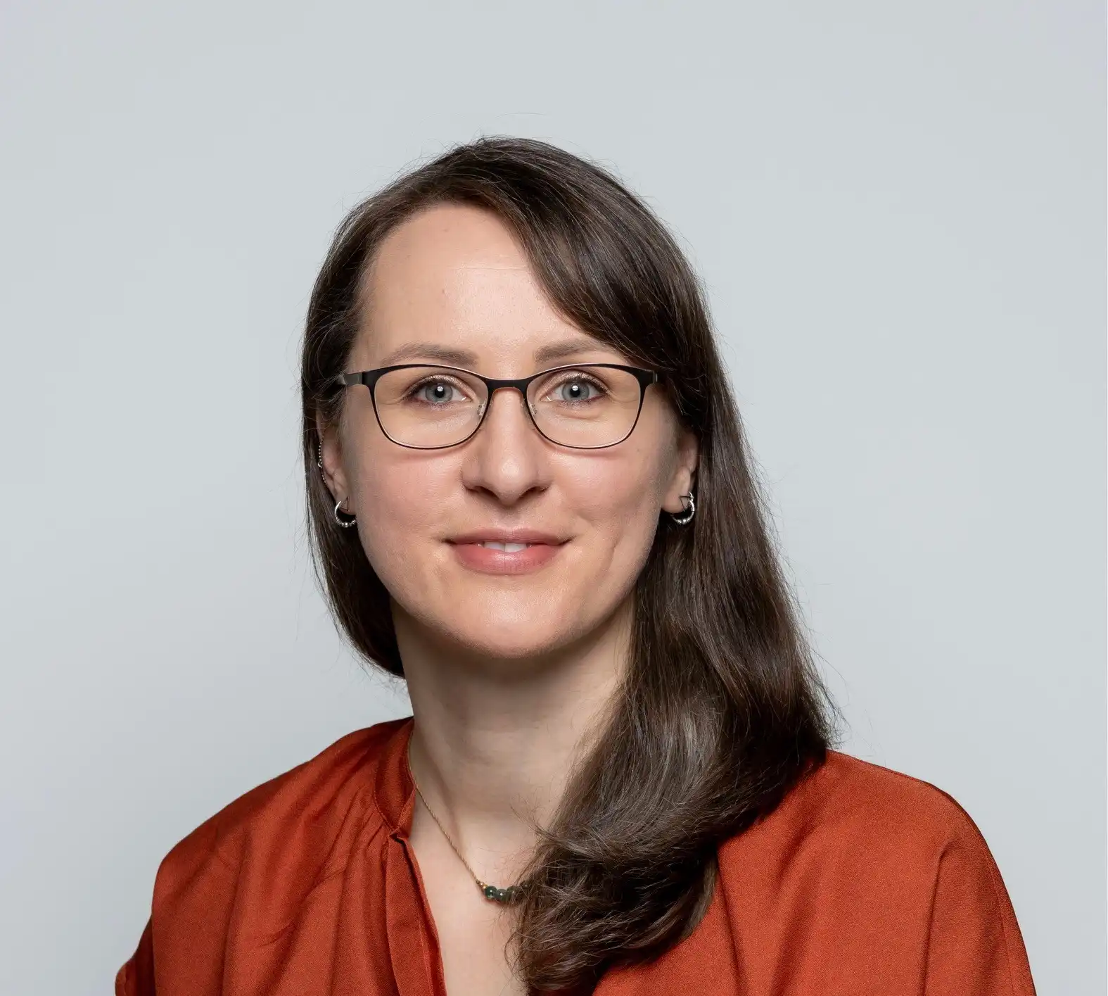 Portrait of a smiling woman with brown hair and glasses, wearing a rust-colored blouse against a gray background.